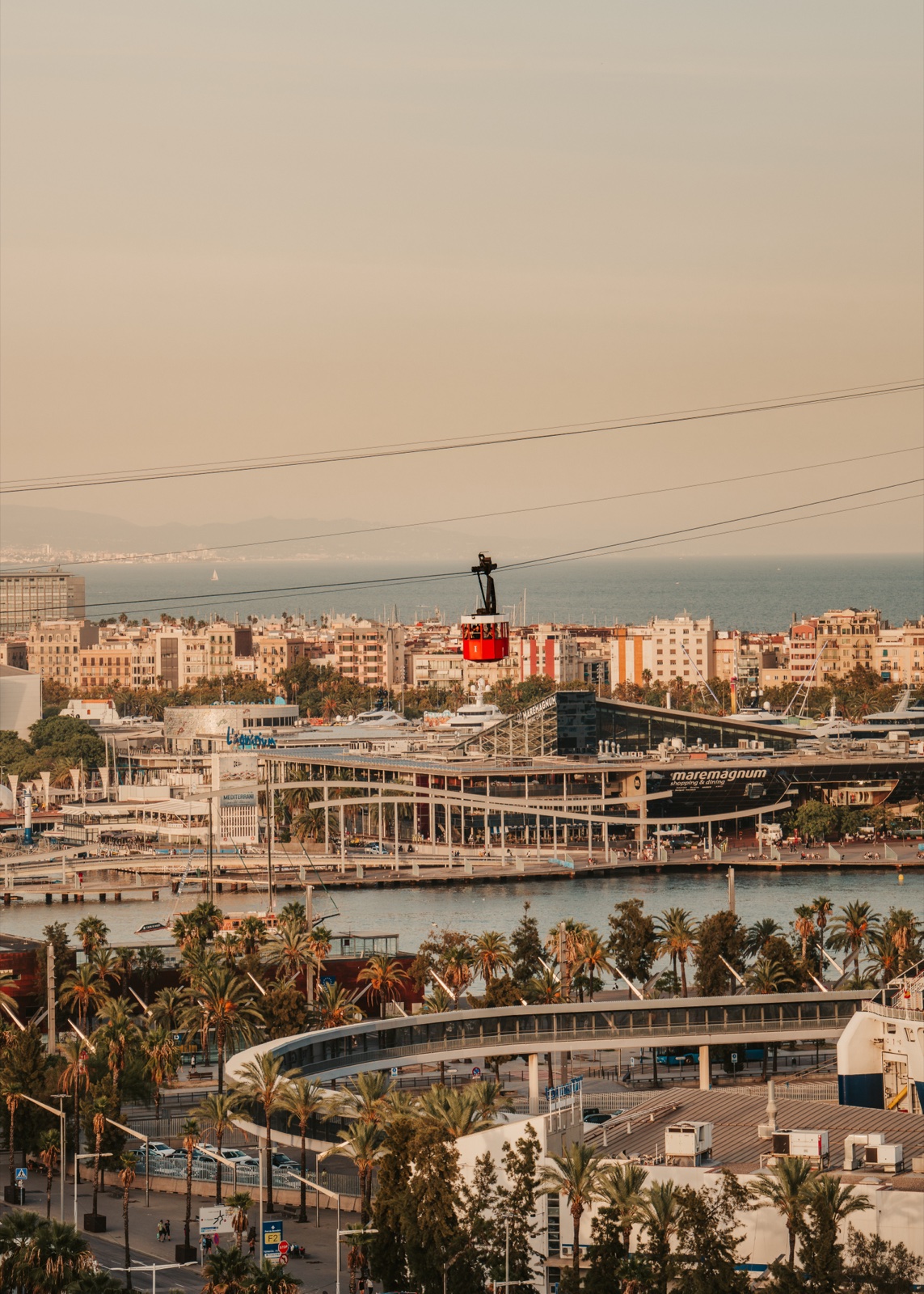 A red cable car crossing above Port Vell and the Maremagnum, with the city and the sea beyond