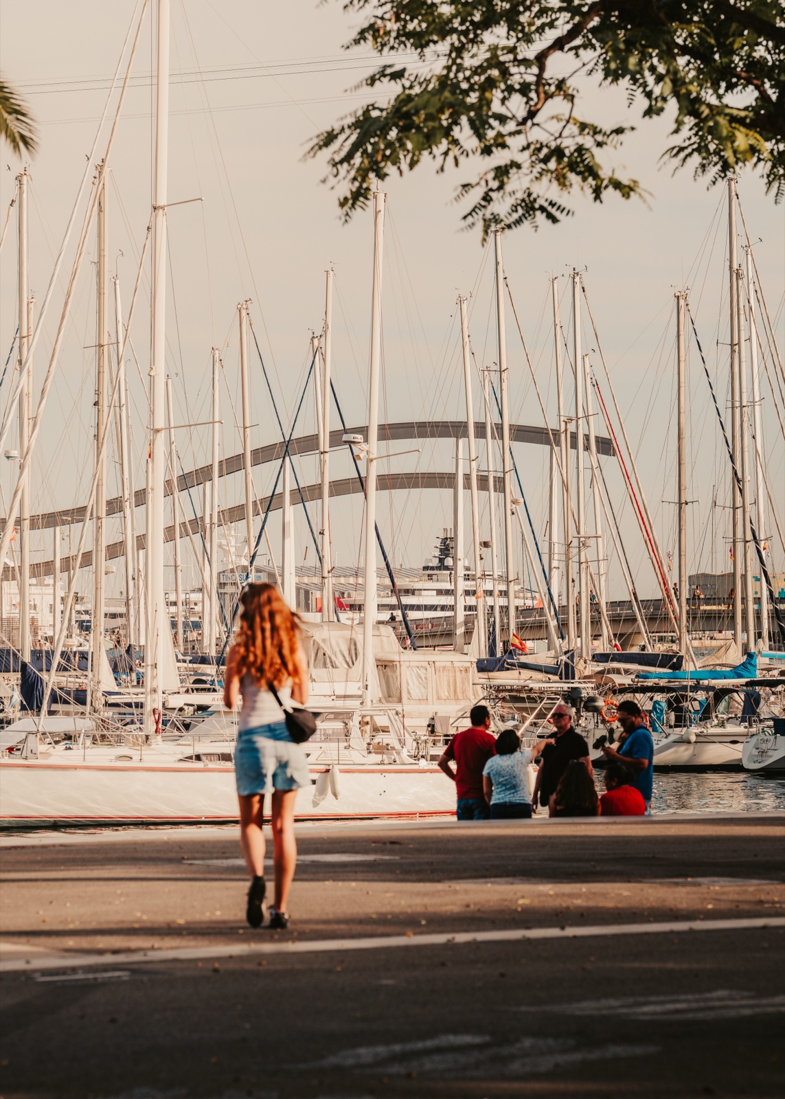 A woman walking towards the moored sailing boats, with people sitting by the water and the footbridge’s arches beyond