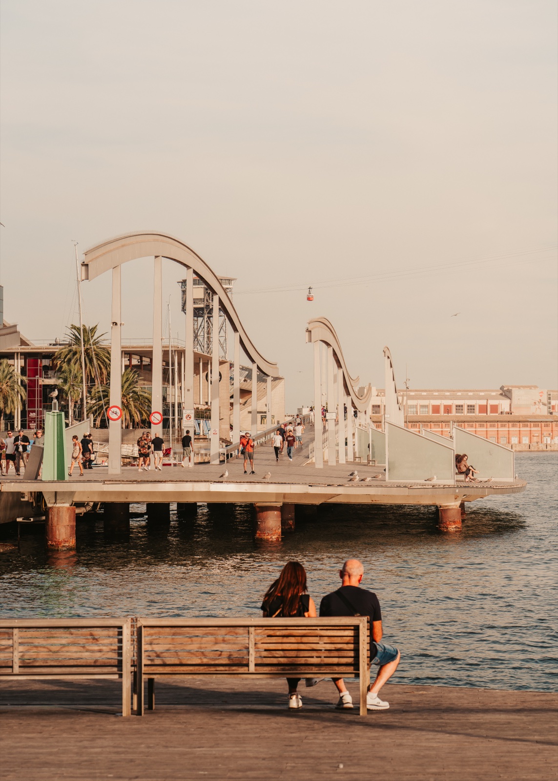 A couple on a bench by the water, looking out at the Rambla de Mar footbridge